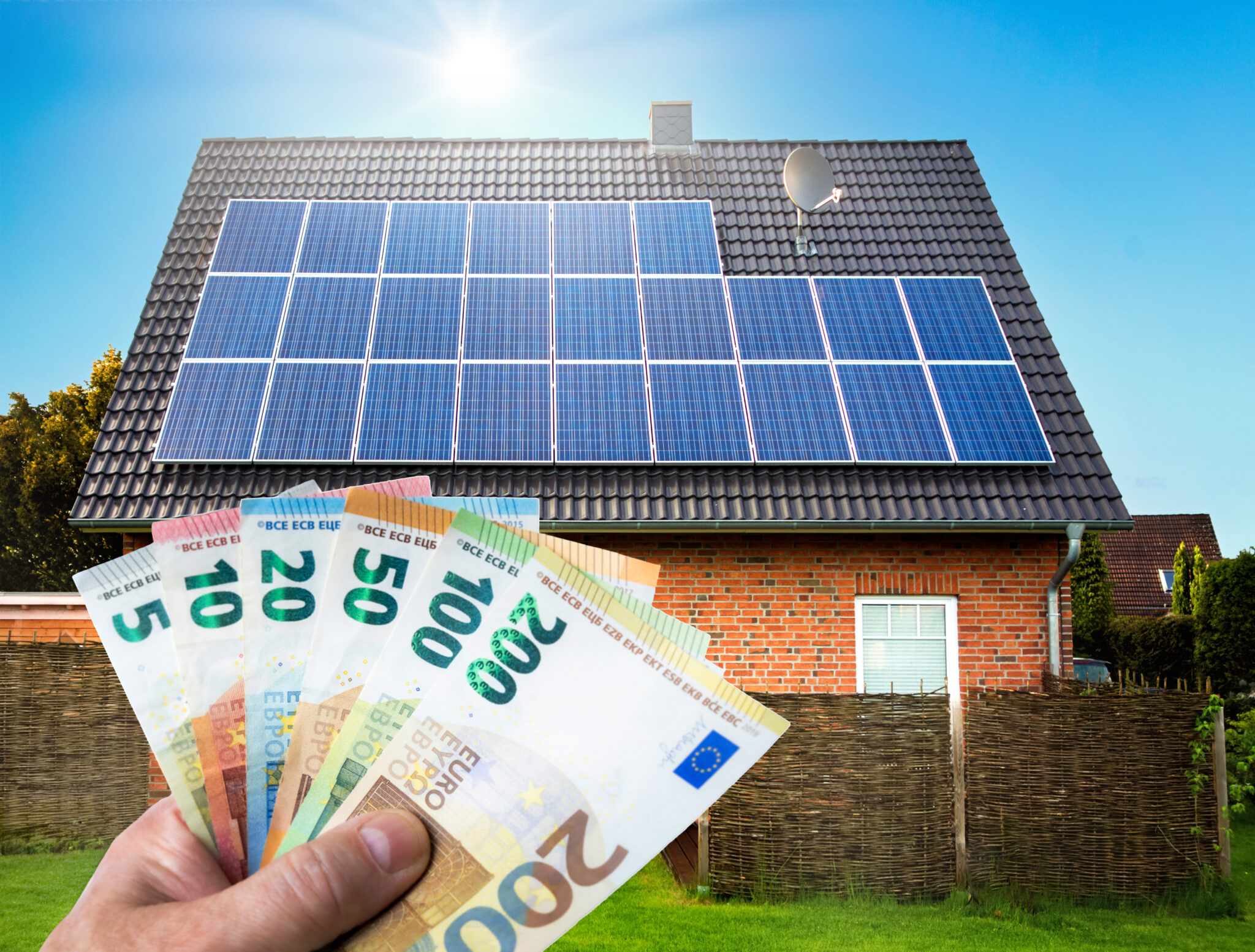 A male hand holds a large sum of European banknotes in front of two single-family houses with solar panels