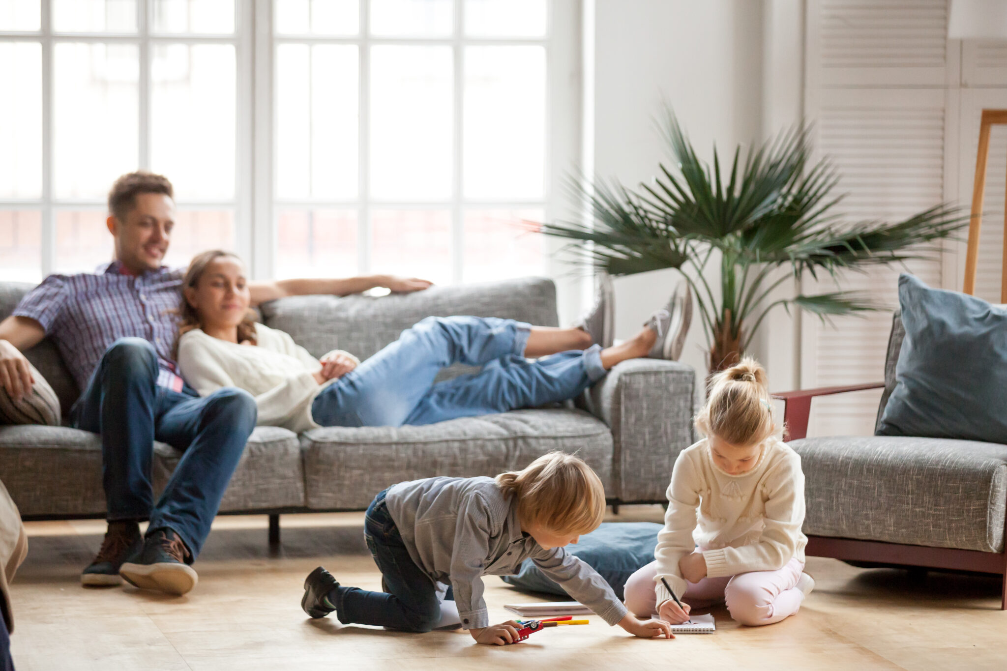 Children sister and brother playing drawing together on floor while young parents relaxing at home on sofa, little boy girl having fun, friendship between siblings, family leisure time in living room